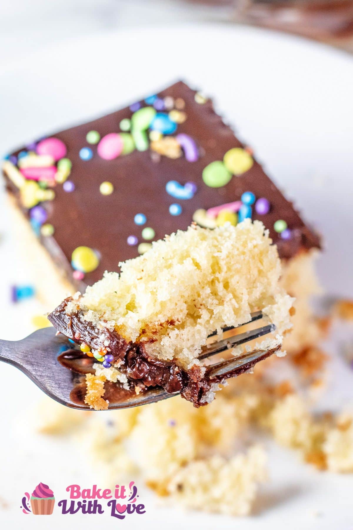 A close-up of a fork holding a bite of yellow cake, revealing its fluffy texture and moist crumbs. The yellow cake is topped with chocolate frosting and colorful sprinkles, with the rest of the slice visible in the blurred background. A small "Bake It With Love" logo is in the bottom left corner.
