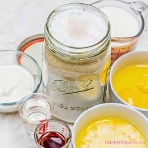 An overhead close-up shows ingredients for yellow cake: a large mason jar filled with layered dry mix (flour, sugar, etc.), and surrounding bowls of wet ingredients like eggs, melted butter, milk, yogurt, oil, and vanilla extract. "BakeItWithLove.com" is at the bottom right.