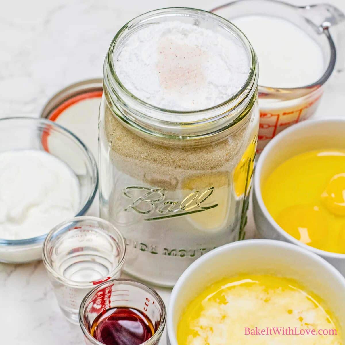 An overhead close-up shows ingredients for yellow cake: a large mason jar filled with layered dry mix (flour, sugar, etc.), and surrounding bowls of wet ingredients like eggs, melted butter, milk, yogurt, oil, and vanilla extract. "BakeItWithLove.com" is at the bottom right.