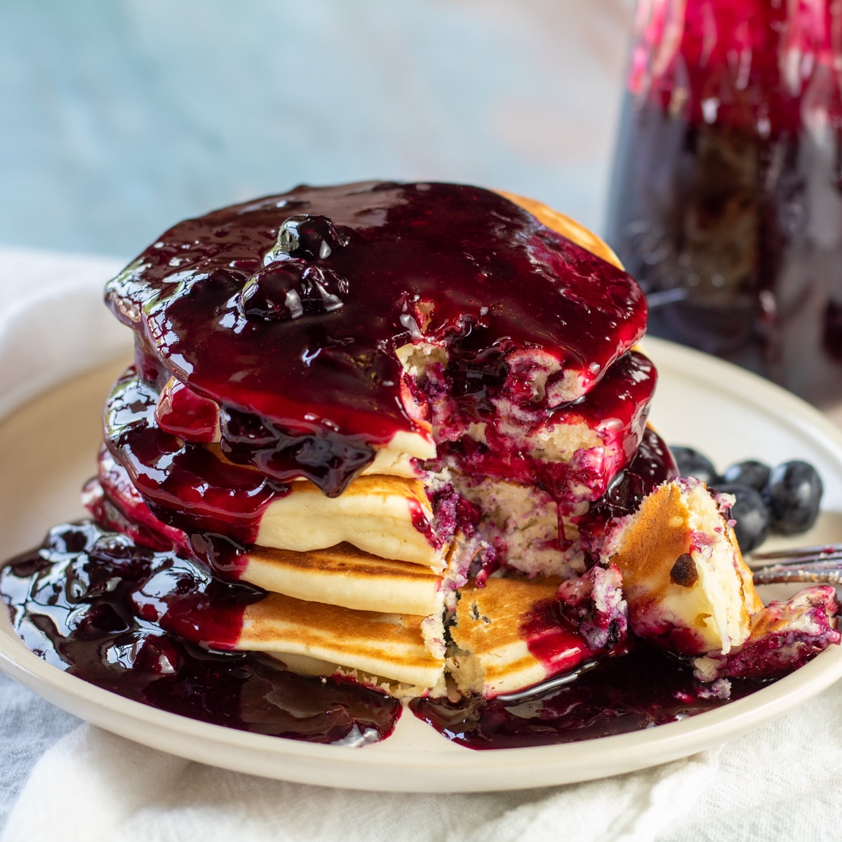 Square image of blueberry pancakes with blueberry syrup on a plate.