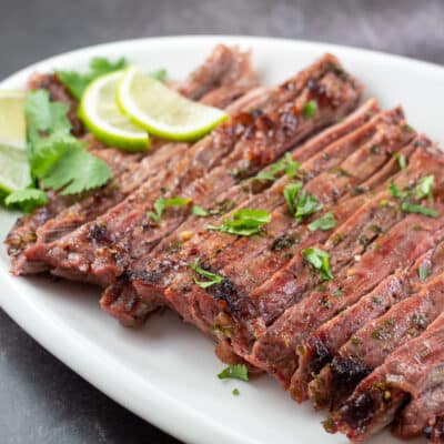 Square image showing carne asada with sliced limes on a white serving platter.