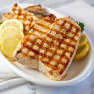 Square image of grilled swordfish on a white plate with fresh lemon slices.