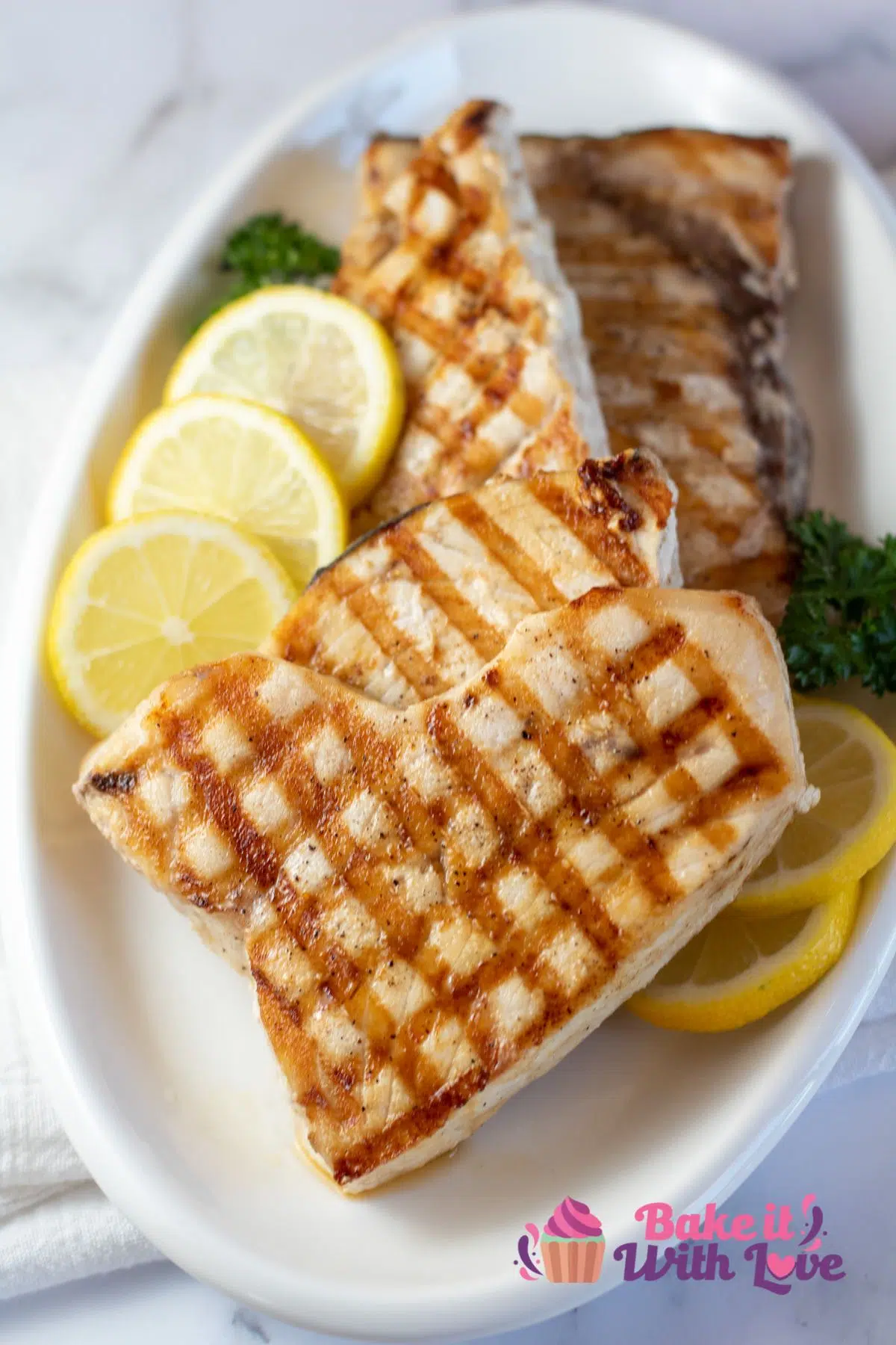 Tall image of grilled swordfish on a white plate with fresh lemon slices.