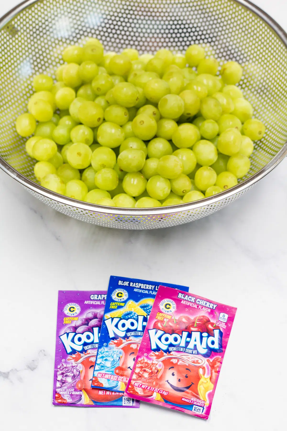 Kool-Aid grapes ingredients with 3 packets of koolaid flavors and a colander with green grapes.