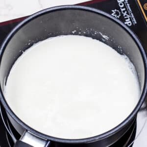 A square image showing a close-up of a wide, shallow saucepan on an electric stovetop filled with thick and creamy scalded milk.