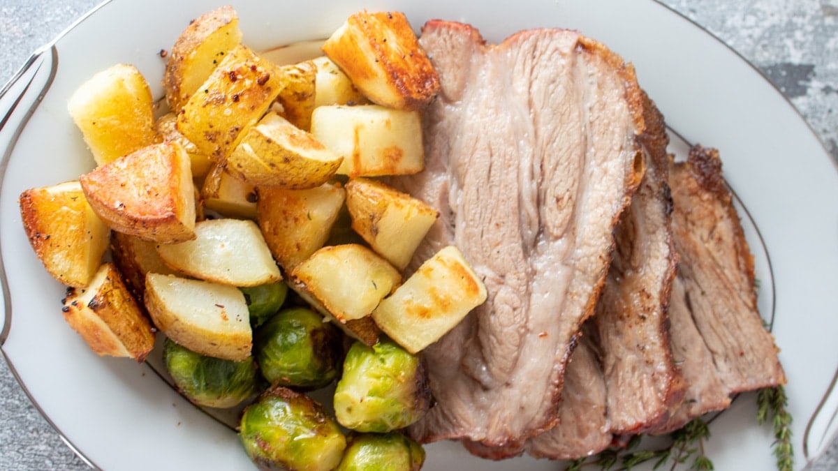 Wide image of pork shoulder butt roast on a plate with potatoes and brussel sprouts.