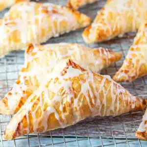 Baked puff pastry apple turnovers with vanilla glaze drizzled over each shown on a wire rack.