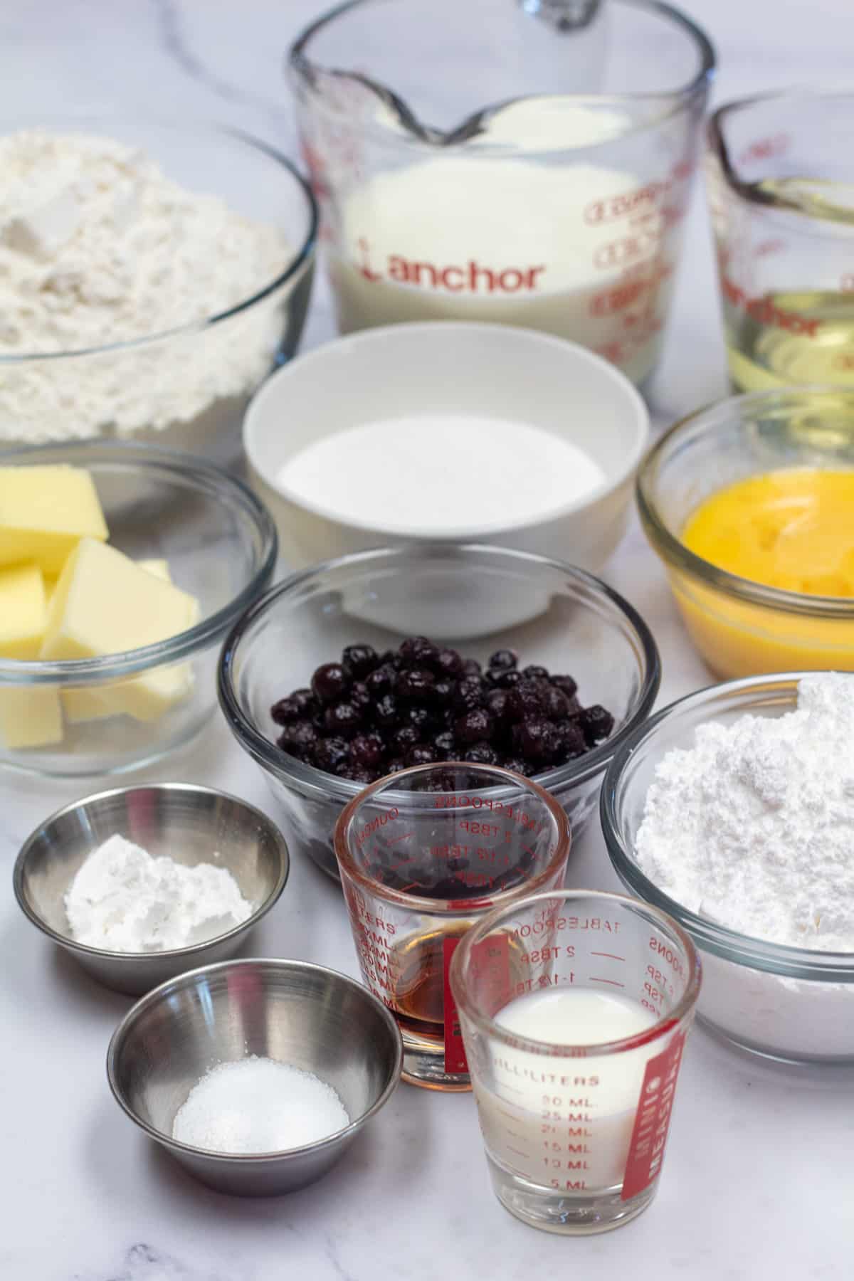Immagine alta che mostra gli ingredienti necessari per preparare le ciambelle ai mirtilli al forno.