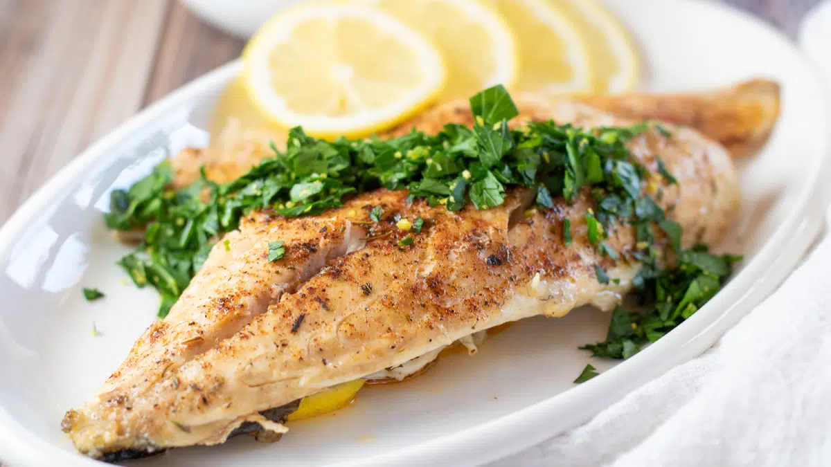 Wide image of branzino with gremolata and lemon slices on a white serving plate.