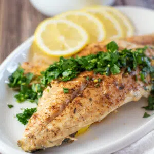 Square image of branzino with gremolata and lemon slices on a white serving plate.