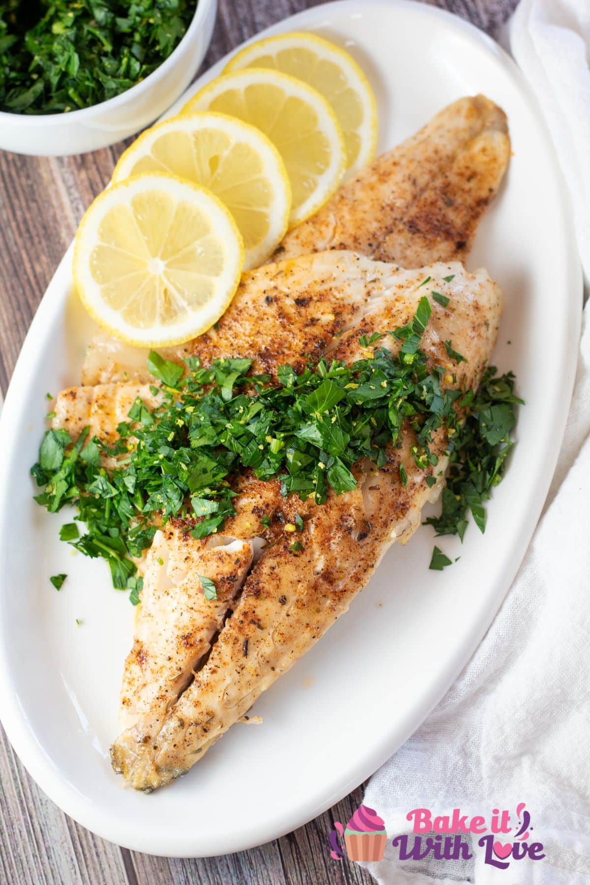 Tall image of branzino with gremolata and lemon slices on a white serving plate.