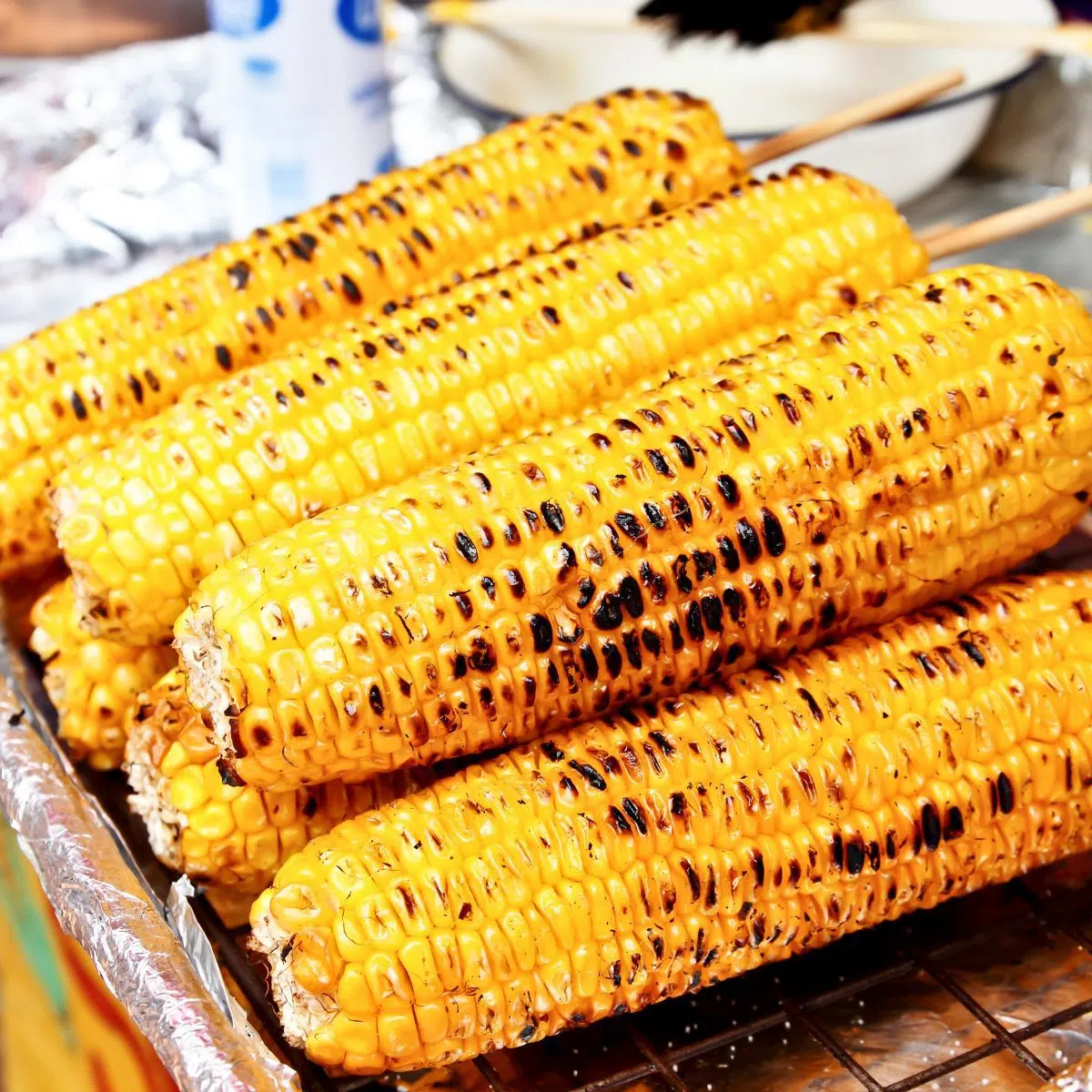 Square image of grilled corn on the cob stacked with wooden skewers.
