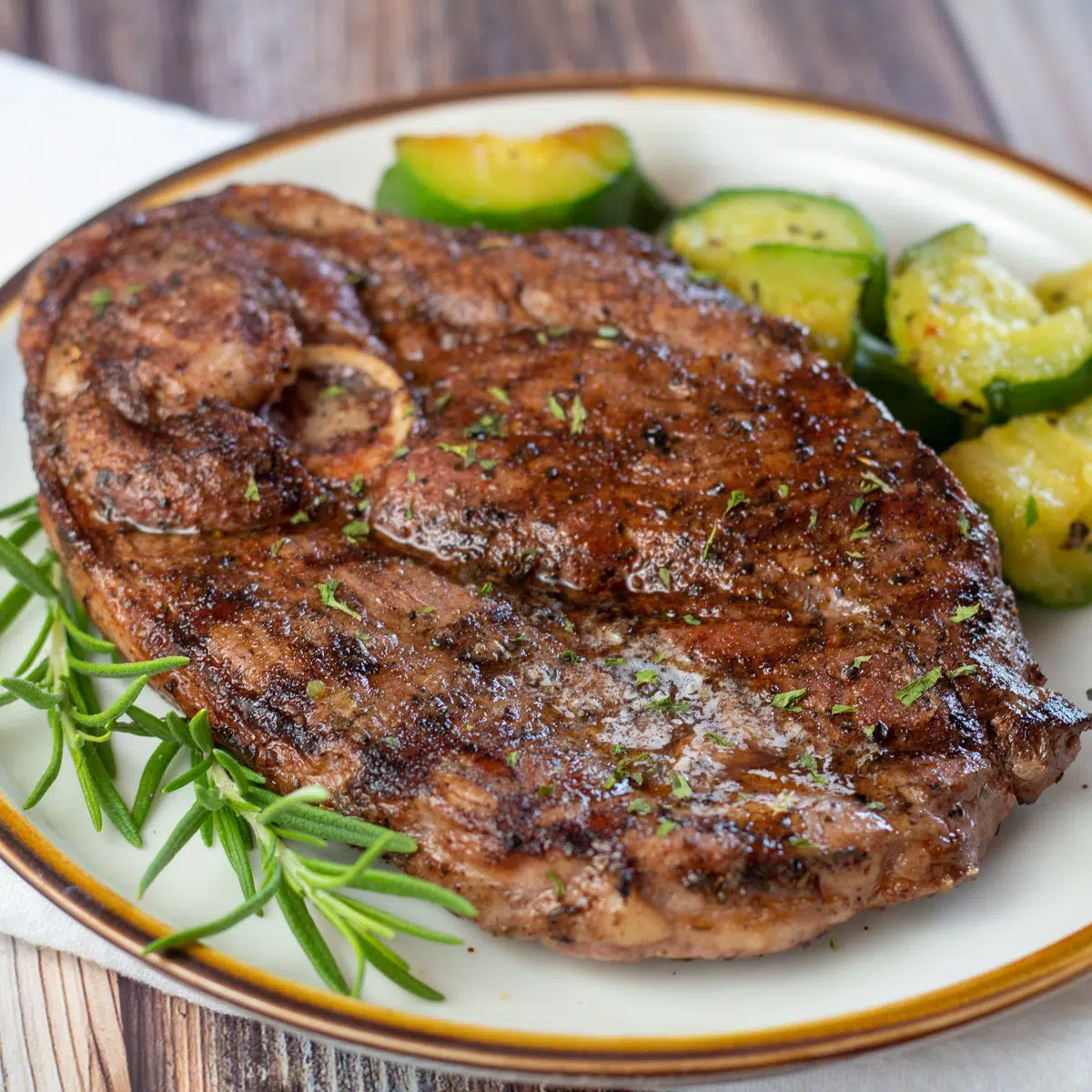 Square image of grilled lamb steak on a plate with zucchini and rosemary.