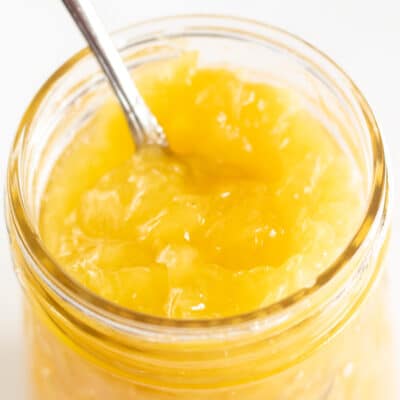 Square image of pineapple pie filling in a glass jar with a spoon inside the jar.