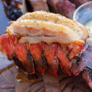 Square image of smoked lobster tail on a wood cutting board with melted butter.