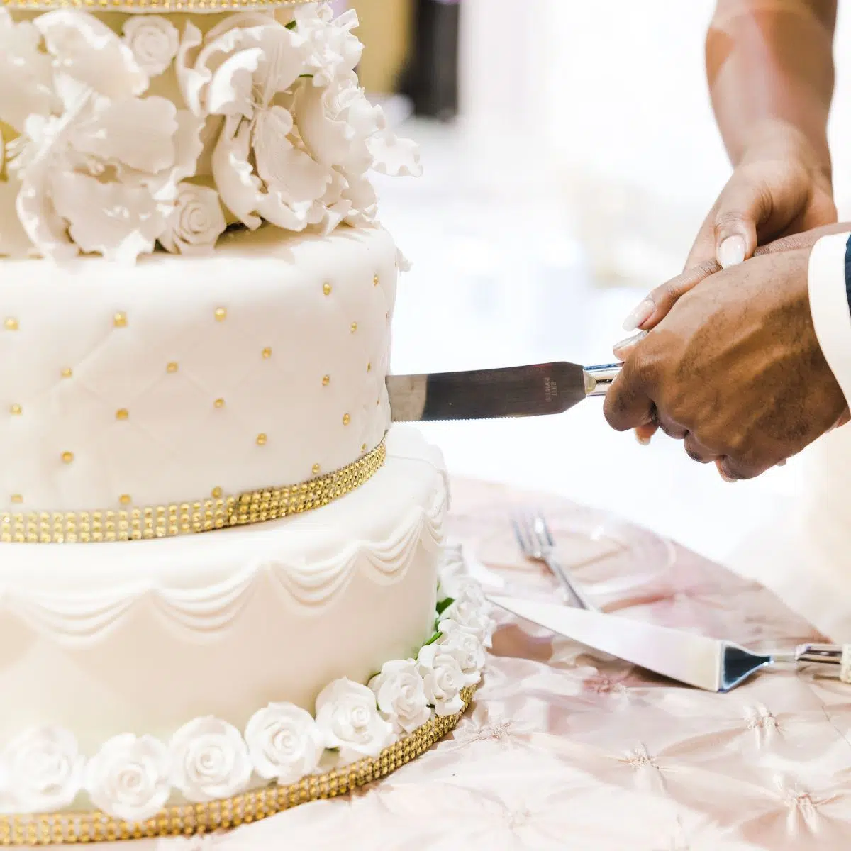 Imagem quadrada de um bolo de casamento sendo cortado.