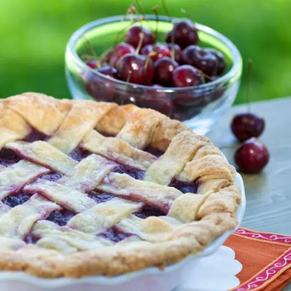 Square image of a cherry pie with cherries in the background.