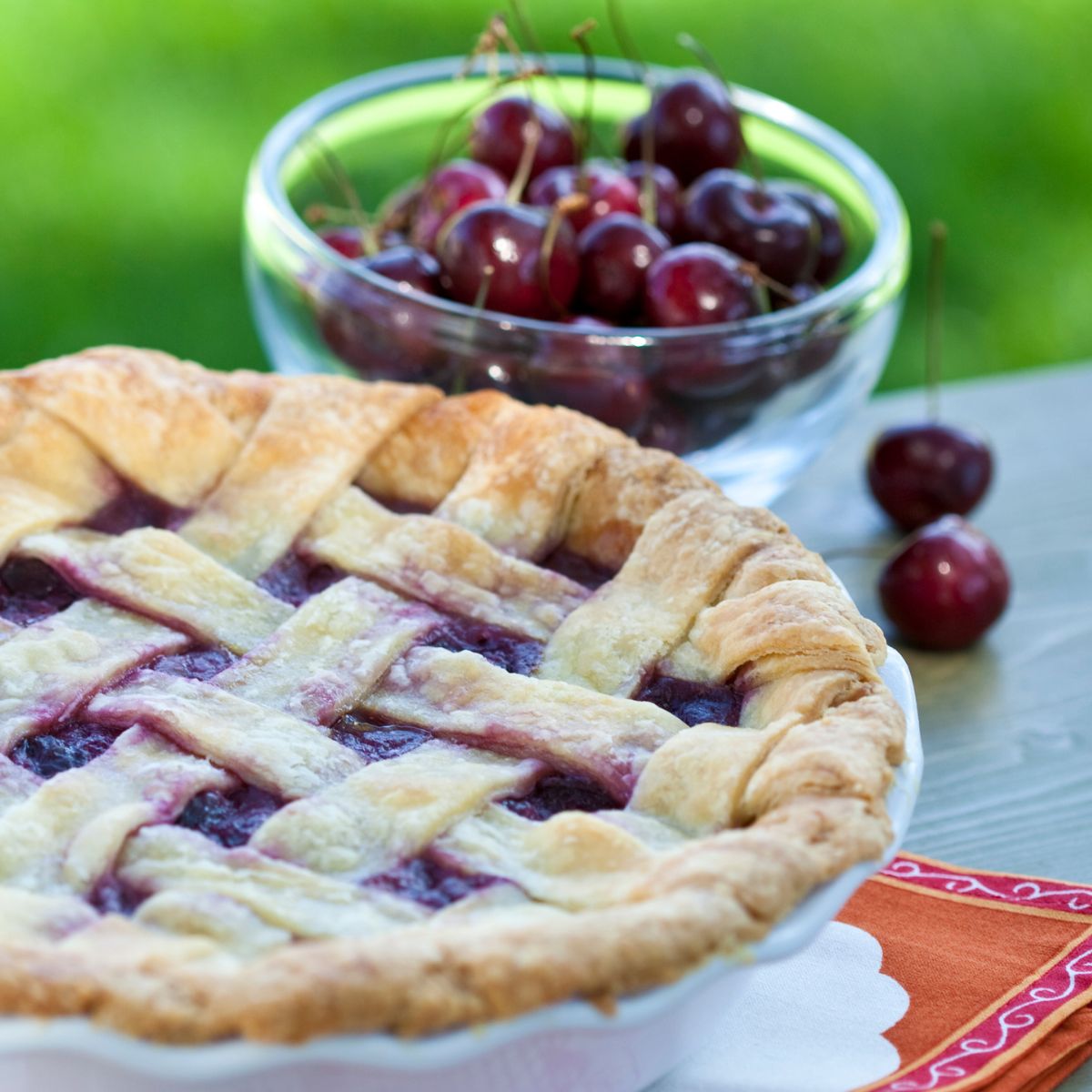 Square image of a cherry pie with cherries in the background.