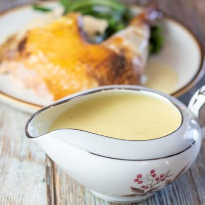 Square image of chicken gravy in a gravy dish next to a plated whole roasted chicken.