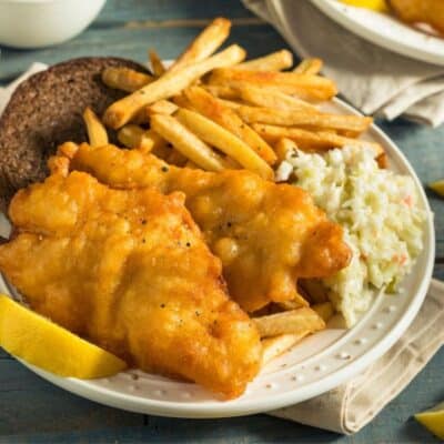Square image of deep fried catfish on a plate with coleslaw and fries.