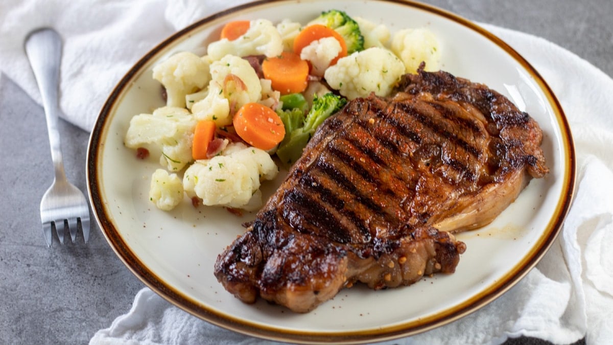 Gambar lebar steak ribeye panggang di piring dengan campuran sayuran.