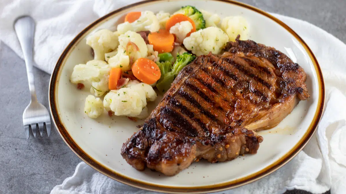 Wide image of a grilled ribeye steak on a plate with mixed veggies.