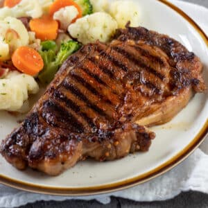 Close up square image of a grilled ribeye steak on a plate with mixed veggies.