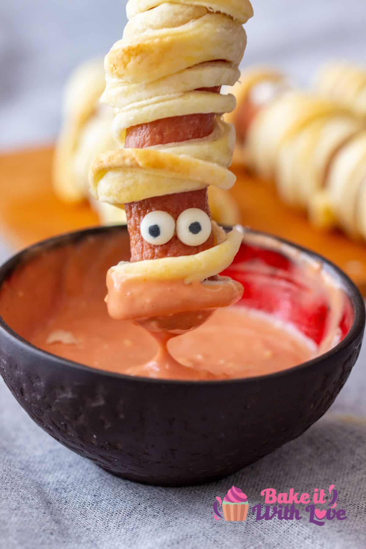 Tall image of "mummy dog" being dipped in sauce.