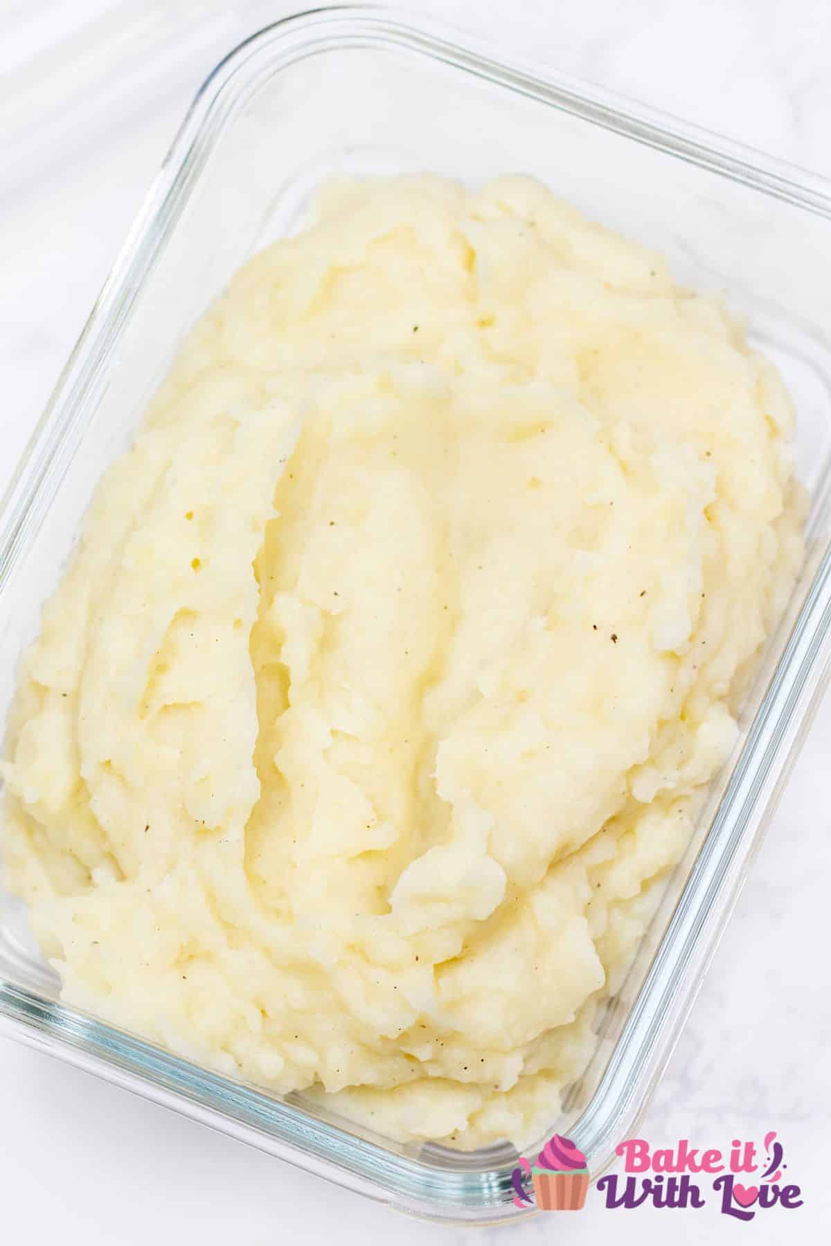 A tal image of creamy mashed potatoes spread evenly in a glass baking dish so that it can be frozen with a 'BakeItWithLove' logo in the bottom right corner.