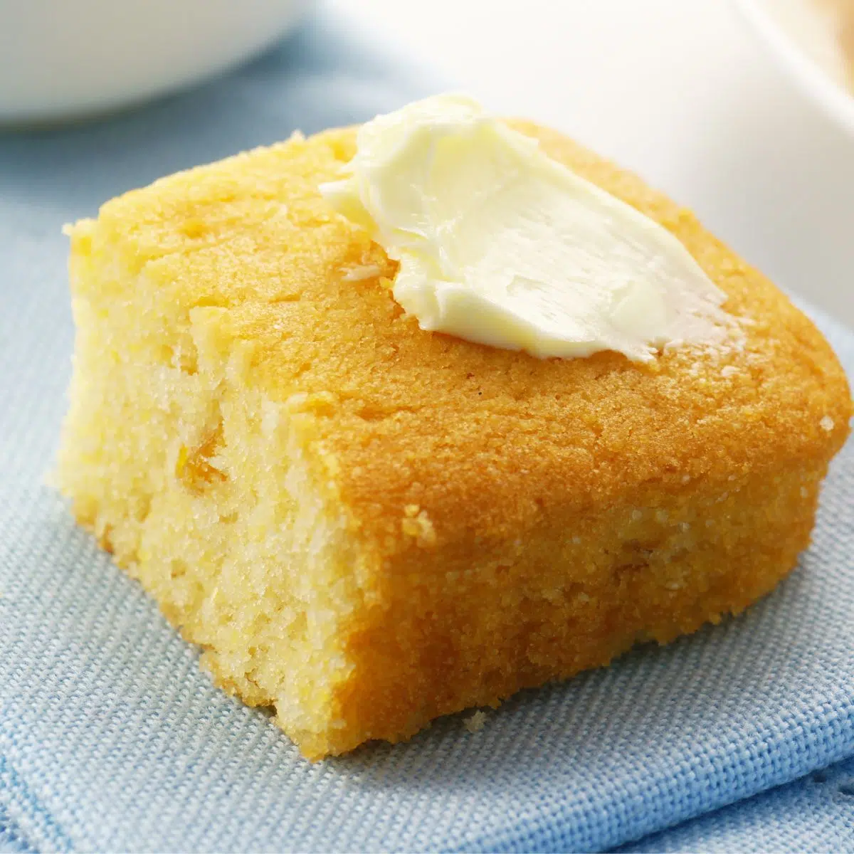 Square image of a piece of cornbread with a pat of butter on top.