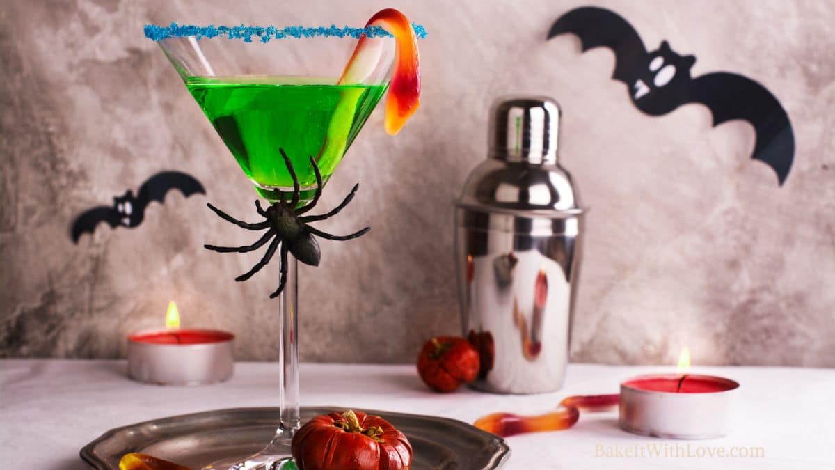 A horizontal image showing a green witch's brew cocktail with blue sugar rim and a gummy snake garnish.