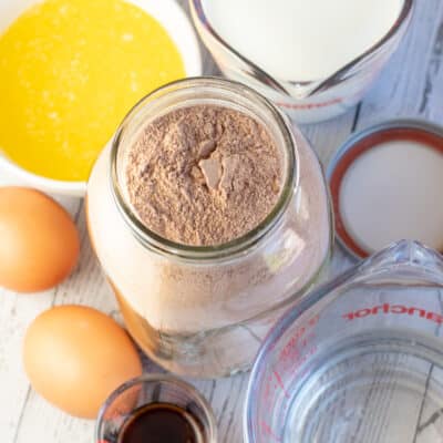 Square image of chocolate cake mix in a mason jar with other cake ingredients around.
