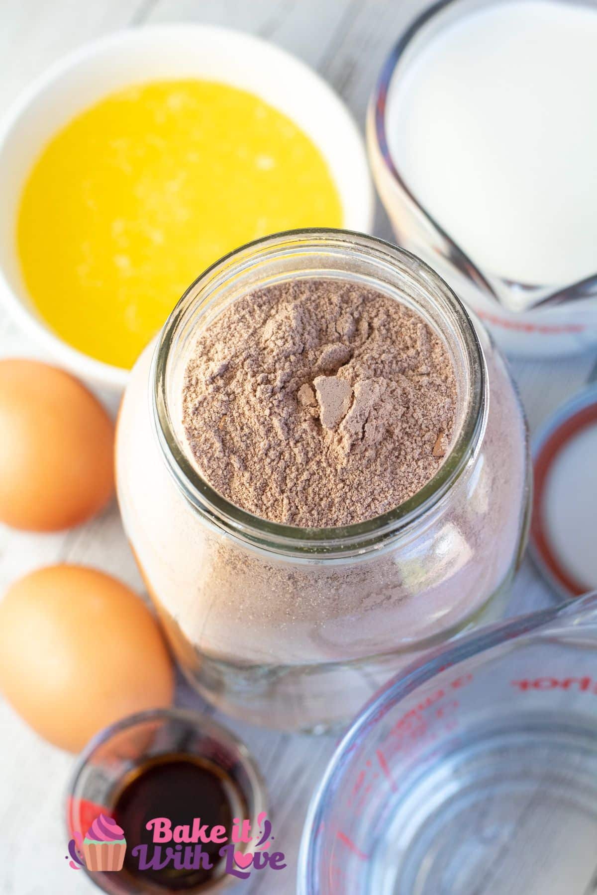 Tall image of chocolate cake mix in a mason jar with other cake ingredients around.