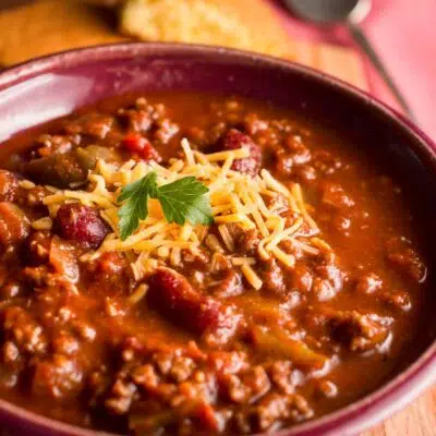 Easy homemade chili recipe served in maroon bowl with shredded cheddar cheese.