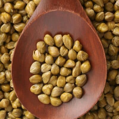 Square image of capers in a wooden spoon.