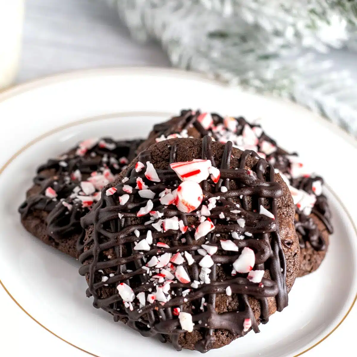 Delicious triple chocolate peppermint cookies on a white plate with festive greenery in the background and a glass of milk.