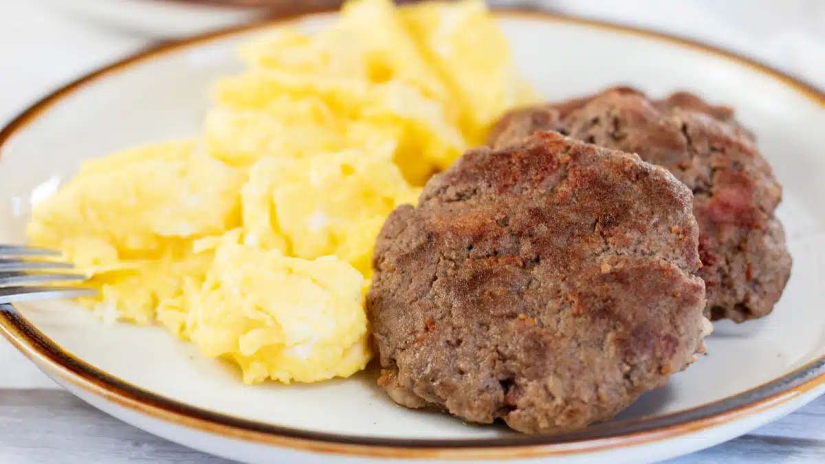 Wide image showing breakfast sausage patties on a plate with scrambled eggs.
