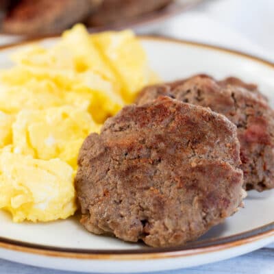 Square image showing breakfast sausage patties on a plate with scrambled eggs.