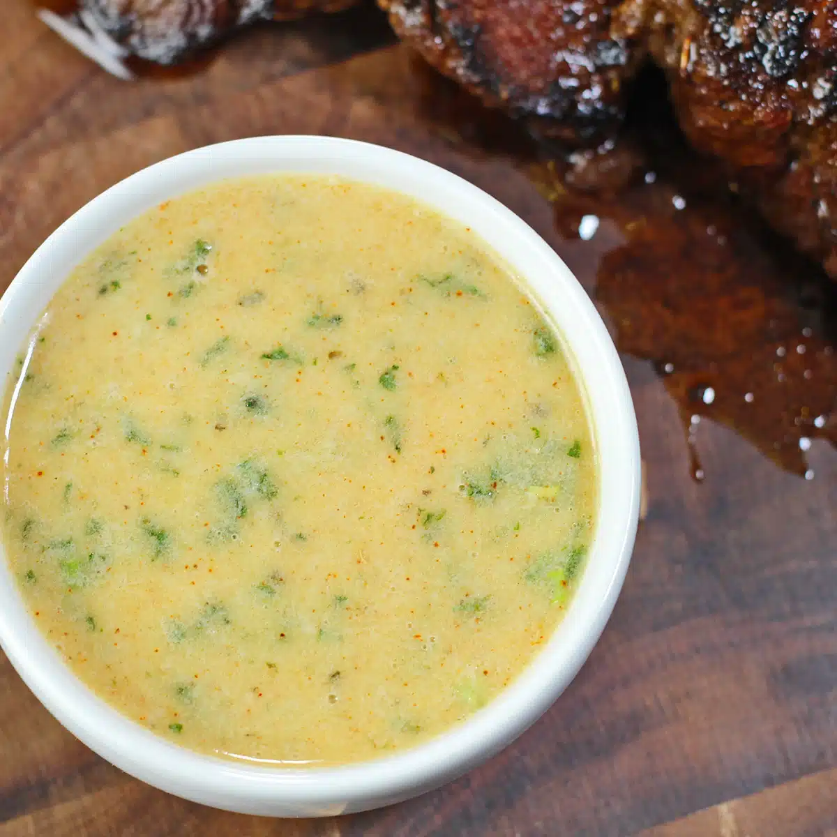 Best cowboy butter recipe with a white sauce bowl filled with the tasty butter dipping sauce and a panseared steak in background.