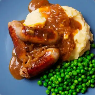 Square image of bangers and mash, onion gravy, and peas on a plate.