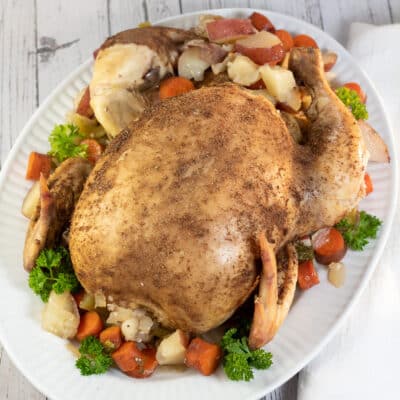 Firkantet billede, der viser en crockpot kogt hel kylling på et serveringsfad med blandede grøntsager.