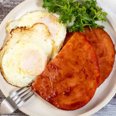 Square image of ham steaks with eggs on a plate.