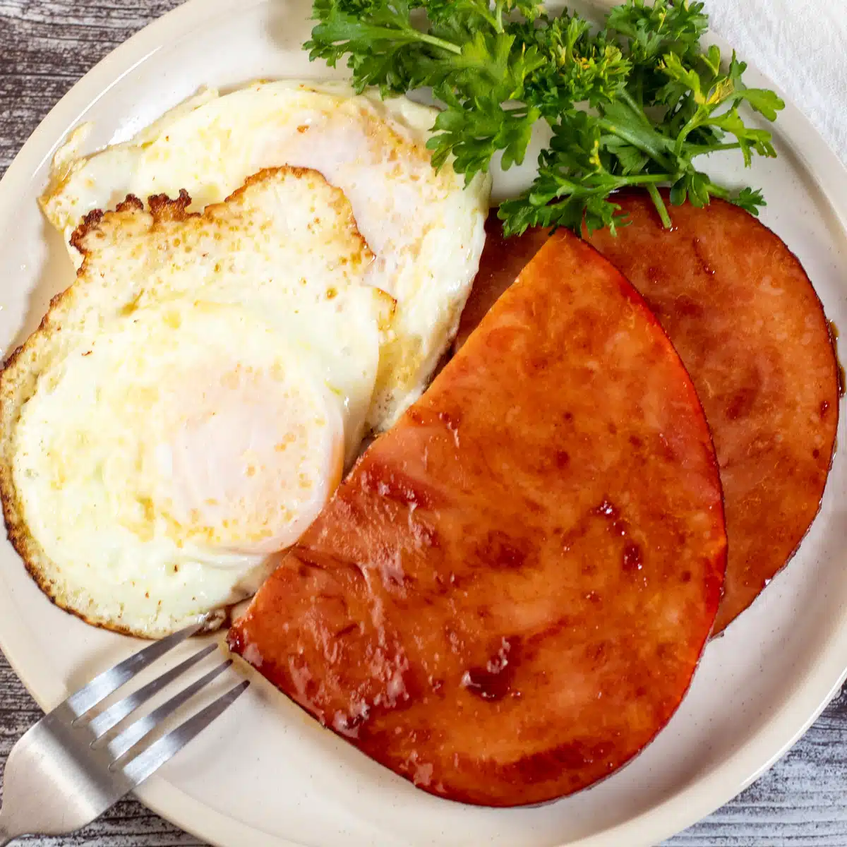 Square image of ham steaks with eggs on a plate.