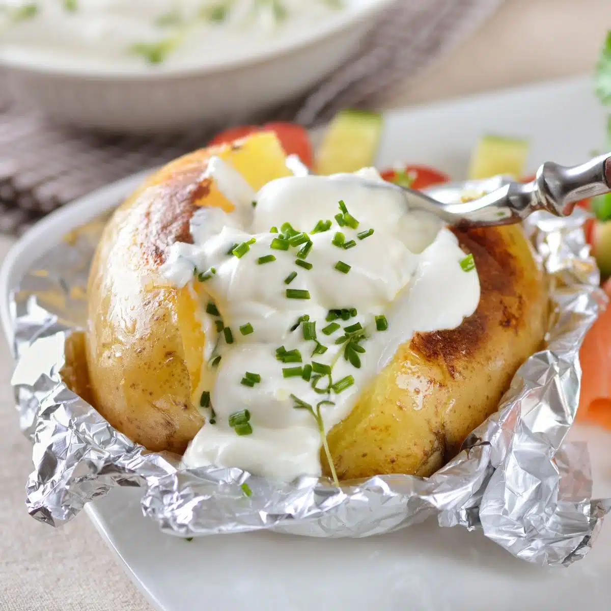 Immagine quadrata che mostra una patata al forno su un piatto bianco, con panna acida ed erba cipollina.