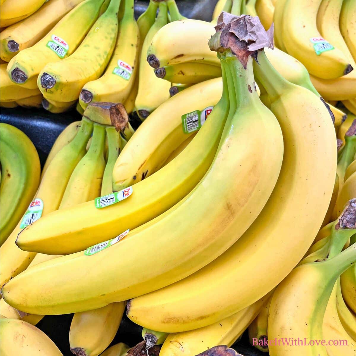 A close-up, square-cropped image of numerous ripe yellow bananas with green stems, piled together in what appears to be a grocery store display. Some bananas have small price or brand stickers on them. The bottom right corner has a faint watermark that says "BakeitWithLove.com"