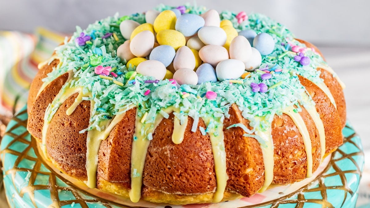 Ampio primo piano sulla mini torta all'uovo di Pasqua con glassa gialla per torta bundt, cocco grattugiato verde, granelli di Pasqua assortiti e mini uova Cadbury nel 'nido'.