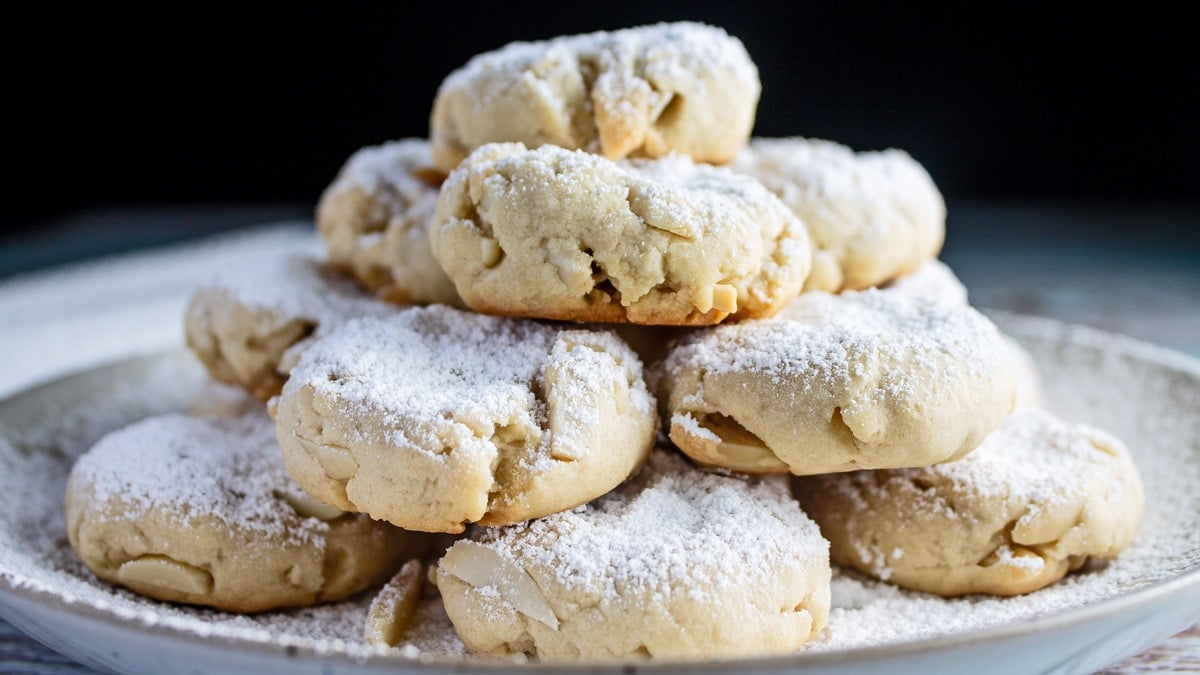Greek almond cookies (kourabiedes) stacked on a plate with generous dusting of confectioners powdered sugar.