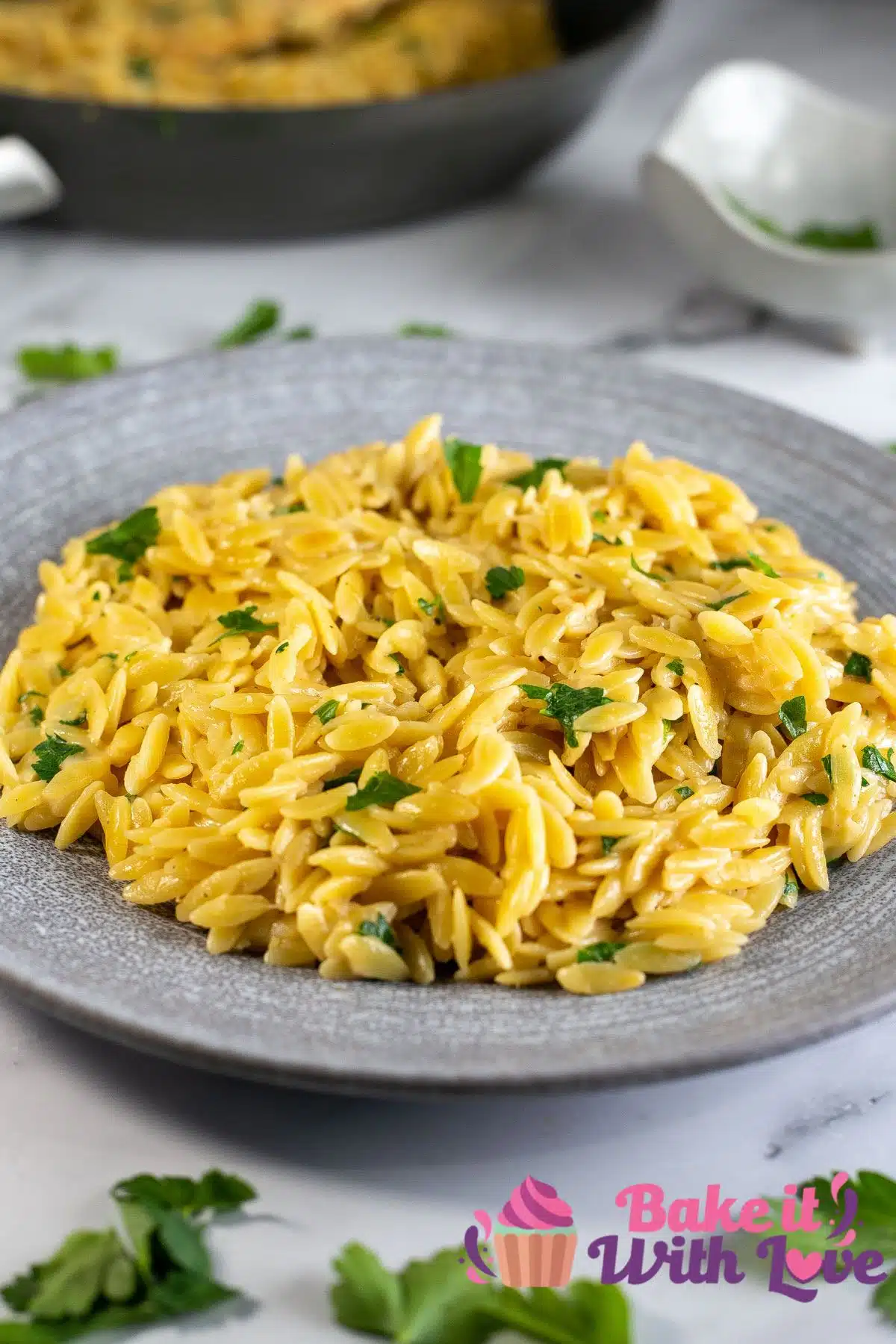 Tasty Parmesan orzo pasta side dish served in grey plate and garnished with fresh chopped parsley with the skillet in background.