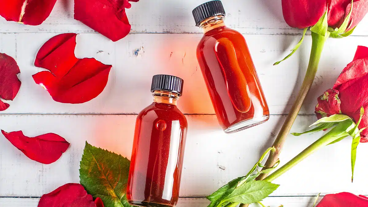 Wide overhead of the DIY rose water in glass bottles on light background with red roses and petals.