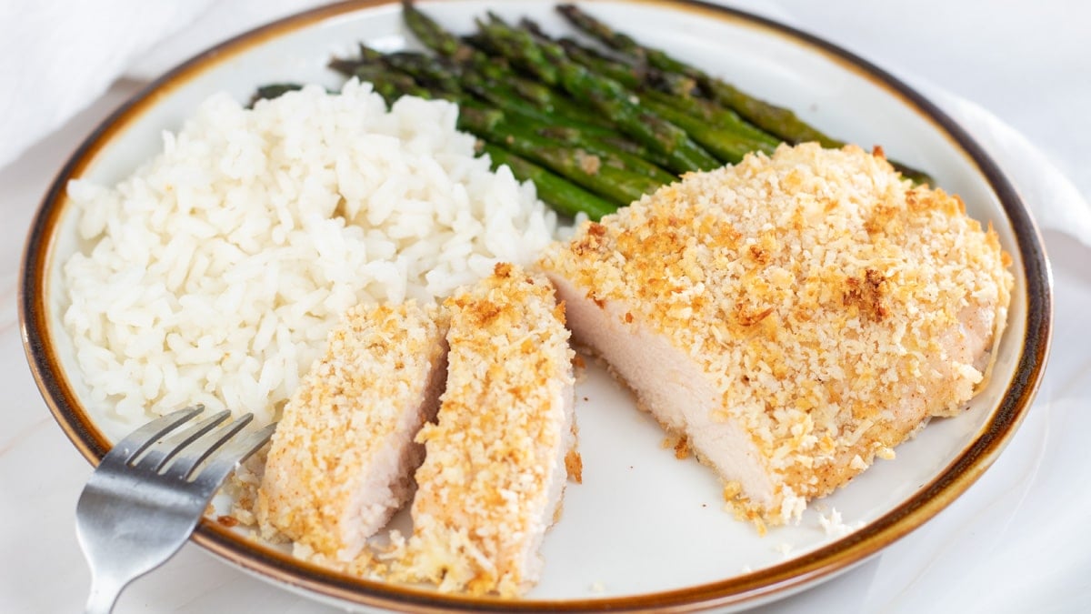 Wide close up image of baked panko chicken on a plate with rice and asparagus.